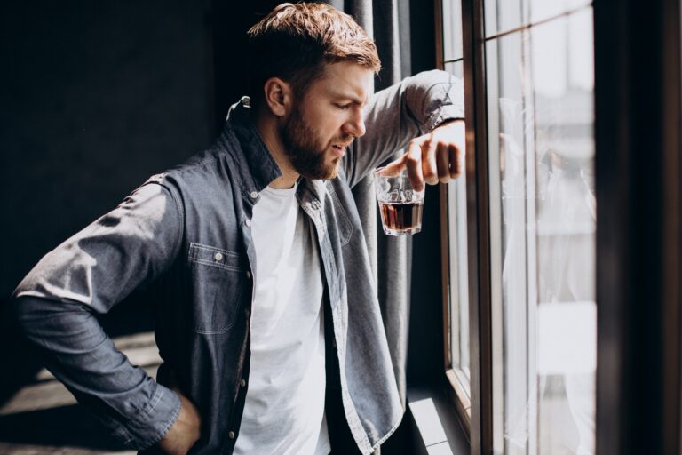 Persona sosteniendo un vaso de agua frente a un fondo desenfocado de botellas de alcohol, simbolizando el desafío de la abstinencia.