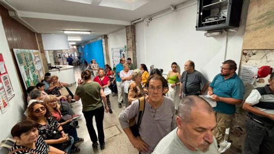 Hombre mayor sentado en silla de ruedas junto a caja de pañales, con un familiar revisando una tablet.