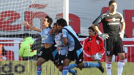 Escudos de Belgrano y River Plate sobre fondo de estadio de fútbol
