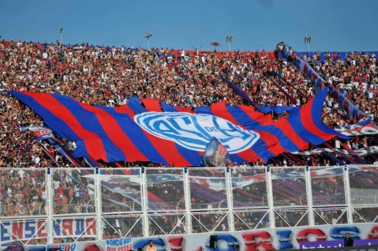 Hinchada de fútbol argentino con banderas y carteles de protesta en un estadio